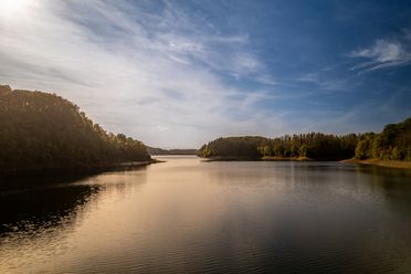 Ein idyllischer See umgeben von bewaldeten Ufern unter blauem Himmel bei Sonnenuntergang.
