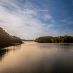 Ein idyllischer See umgeben von bewaldeten Ufern unter blauem Himmel bei Sonnenuntergang.