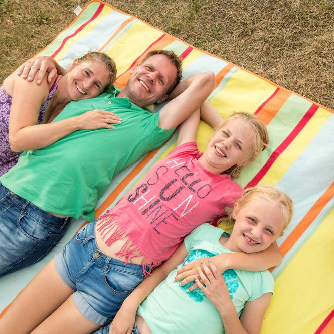 Familie liegt lächelnd auf bunter Decke im Gras; Mutter, Vater und zwei Töchter genießen entspannte Zeit.