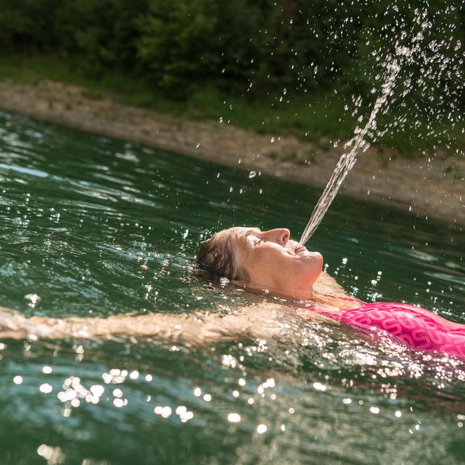 Frau in rosa Badeanzug genießt Wasserstrahl in sonnigem See, mit Bäumen im Hintergrund.