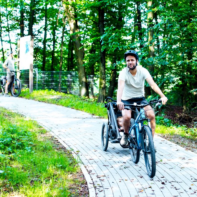 Ein Mann fährt auf einem Fahrrad mit einer Hilfsperson auf einem Waldweg, umgeben von grünen Bäumen.