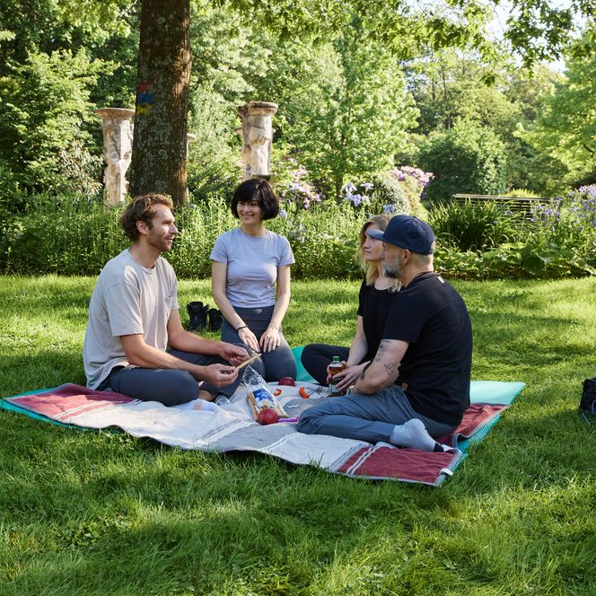 Vier Personen sitzen entspannt auf einer Picknickdecke in einem grünen, blühenden Garten.