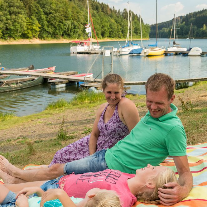 <p>Eine Familie entspannt auf einer bunten Decke am Ufer eines Sees mit Segelbooten.</p>