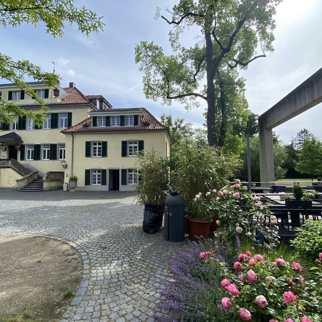 Altes Gebäude mit Ziegeldach, umgeben von Blumen, Bäumen und einem gepflasterten Hof.
