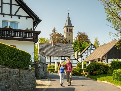 Ein junges Paar wandert auf einer Straße, im Hintergrund stehen Fachwerkhäuser, hinter denen ein Kirchturm emporragt.