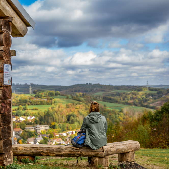 <p>Person sitzt auf Bank vor Holzhütte mit Blick auf eine hügelige, bewaldete Landschaft unter bewölktem Himmel.</p>