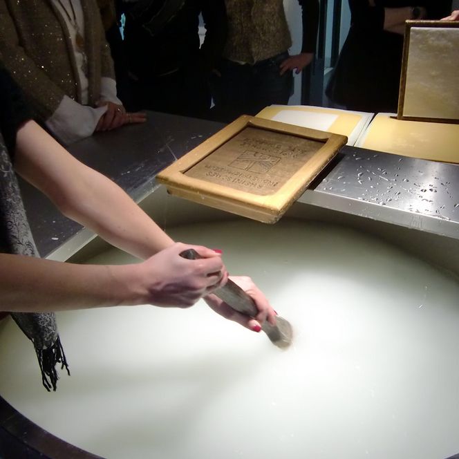 Papierherstellung: Hände schöpfen Papier aus einer Wasserwanne, umgeben von Zuschauern.