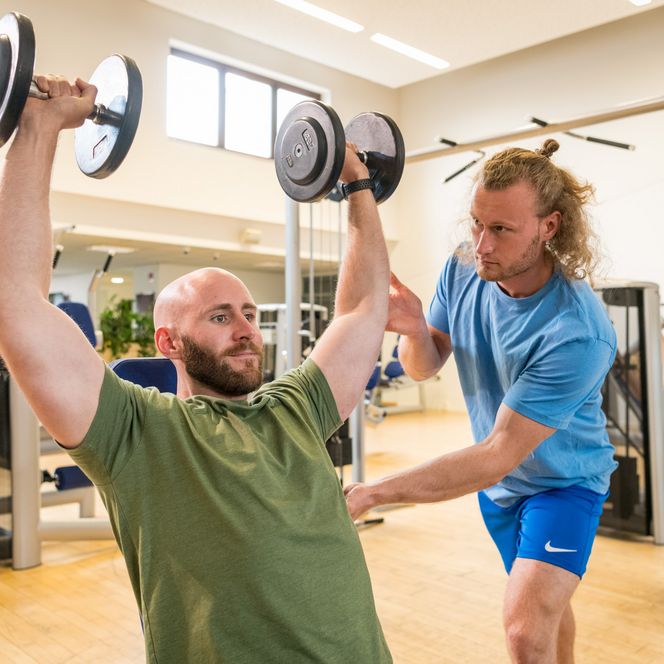 <p>Ein Mann hebt Hanteln im Fitnessstudio, während ein Trainer ihm hilft.</p>