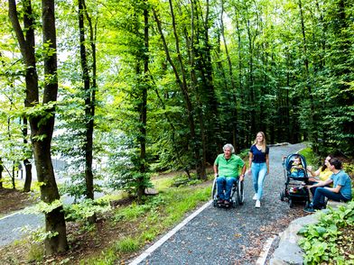 Ein älterer Mann im Rollstuhl und eine Frau spazieren einen Waldweg entlang, gefolgt von einer Mutter mit Kinderwagen.