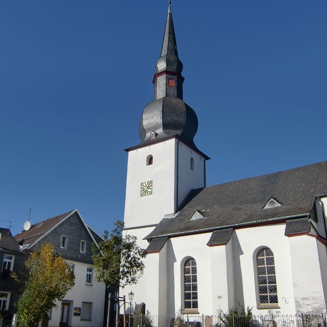 <p>Kirche mit Spitzdach und Fachwerkhäusern im Hintergrund unter blauem Himmel in Bergneustadt.</p>