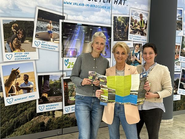 Drei Frauen stehen vor einer beklebten Schaufensterfront eines leeren Ladenlokals in einem Einkaufszentrum. Sie halten gemeinsam einen Flyer in der Hand.