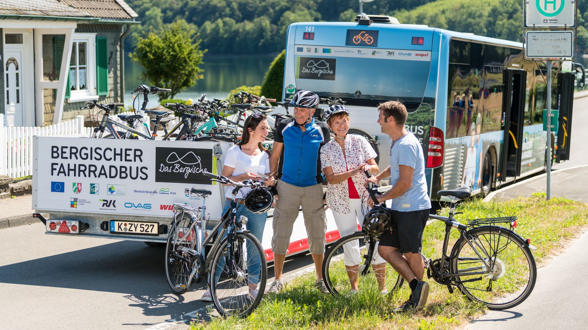 Zwei Männer und zwei Frauen stehen vor dem Anhänger vom Bergischen Fahrradbus. Neben ihnen stehen zwei Fahrräder. Im Hintergrund ist eine Talsperre zu sehen.