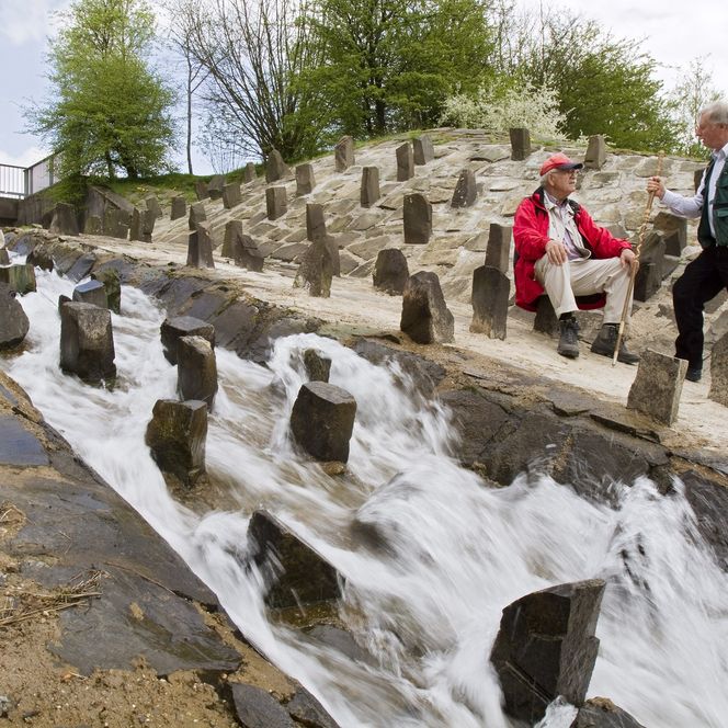 Zwei Männer sitzen neben einer fließenden Wasserrinne mit geometrischen Steinen in naturnaher Umgebung.
