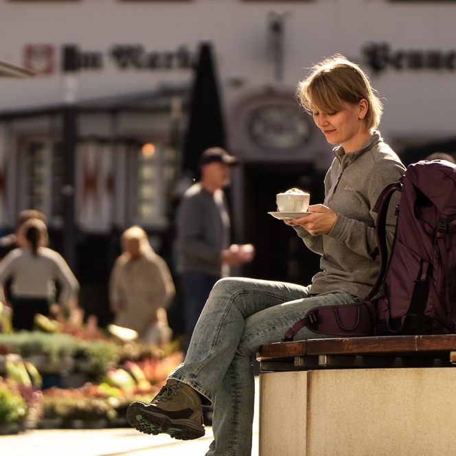 <p>Frau genießt Kaffee auf einer Bank bei sonnigem Wetter in belebter Straßenatmosphäre.</p>