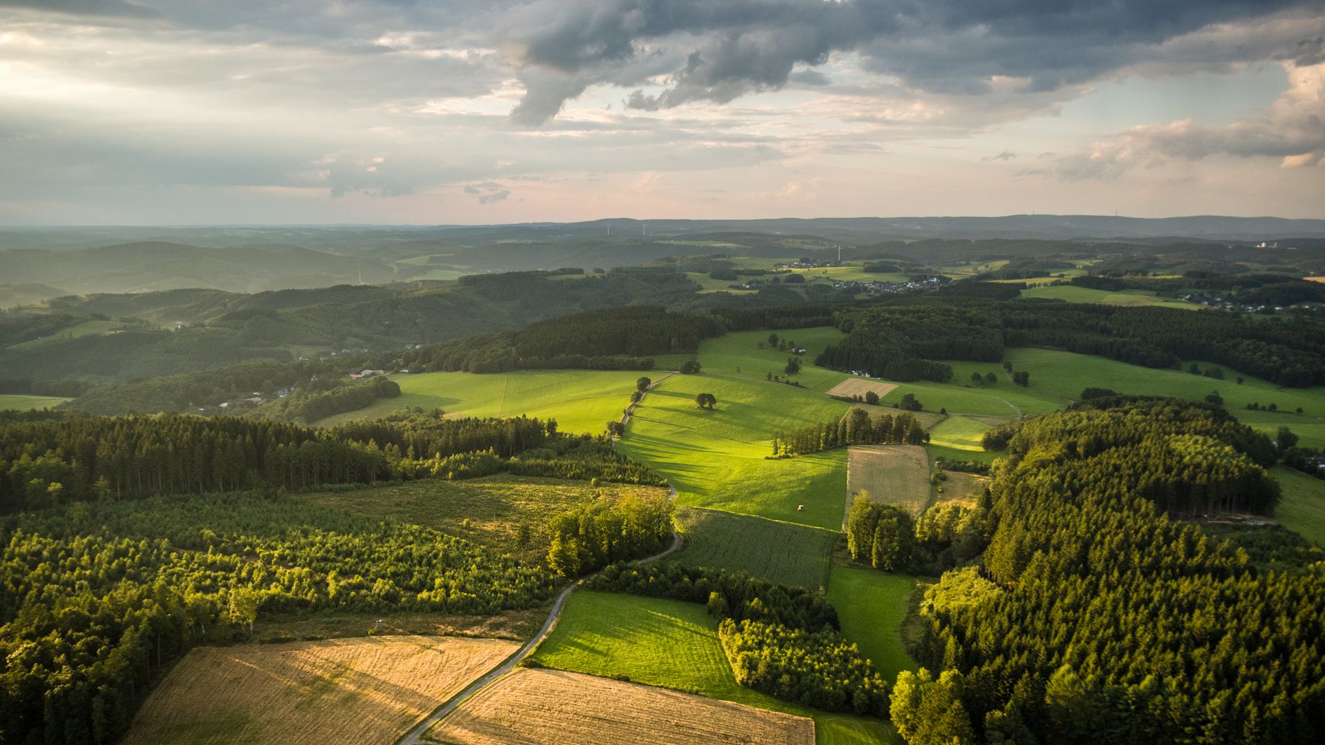 Luftaufnahme von Feldern, Wiesen und Wäldern. Die Sonne scheint durch dunkle Wolken