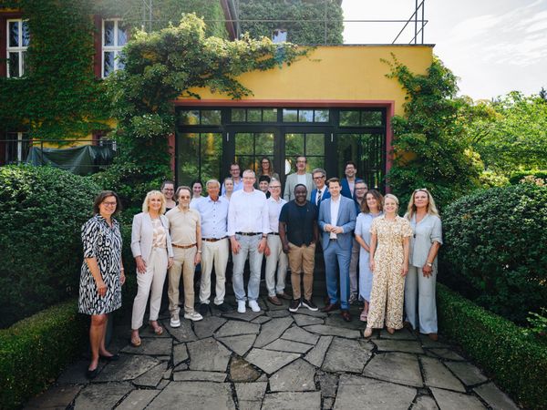 Gruppe von rund 18 Personen bei einem Empfang vor dem Eingang eines gelben, mit Efeu bewachsenen Gebäudes, umgeben von Hecken und Gartenanlage.