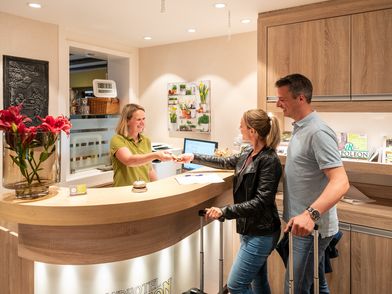 Ein Mann und eine Frau stehen an einer Rezeption in einem Hotel und checken ein. Hinter der Theke steht eine Frau und reicht einen Schlüssel an.