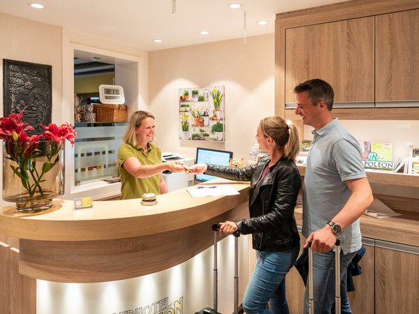 Ein Mann und eine Frau stehen an einer Rezeption in einem Hotel und checken ein. Hinter der Theke steht eine Frau und reicht einen Schlüssel an.