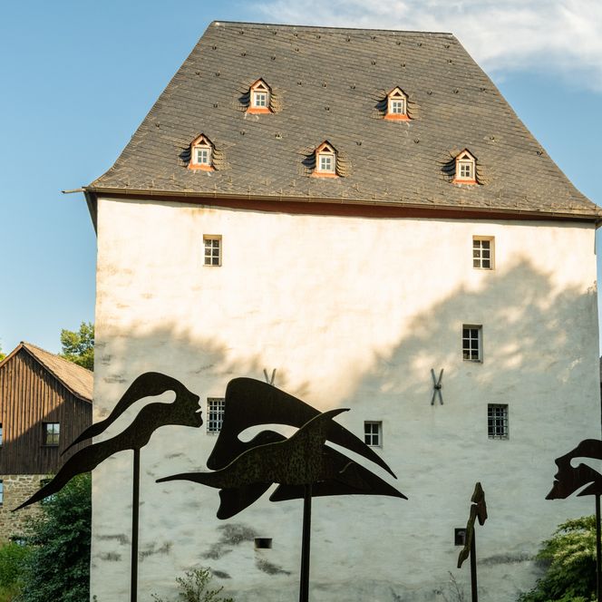 Altes Fachwerkhaus mit Schieferdach, moderne Skulpturen im Vordergrund, umgeben von Bäumen bei Sonnenschein.