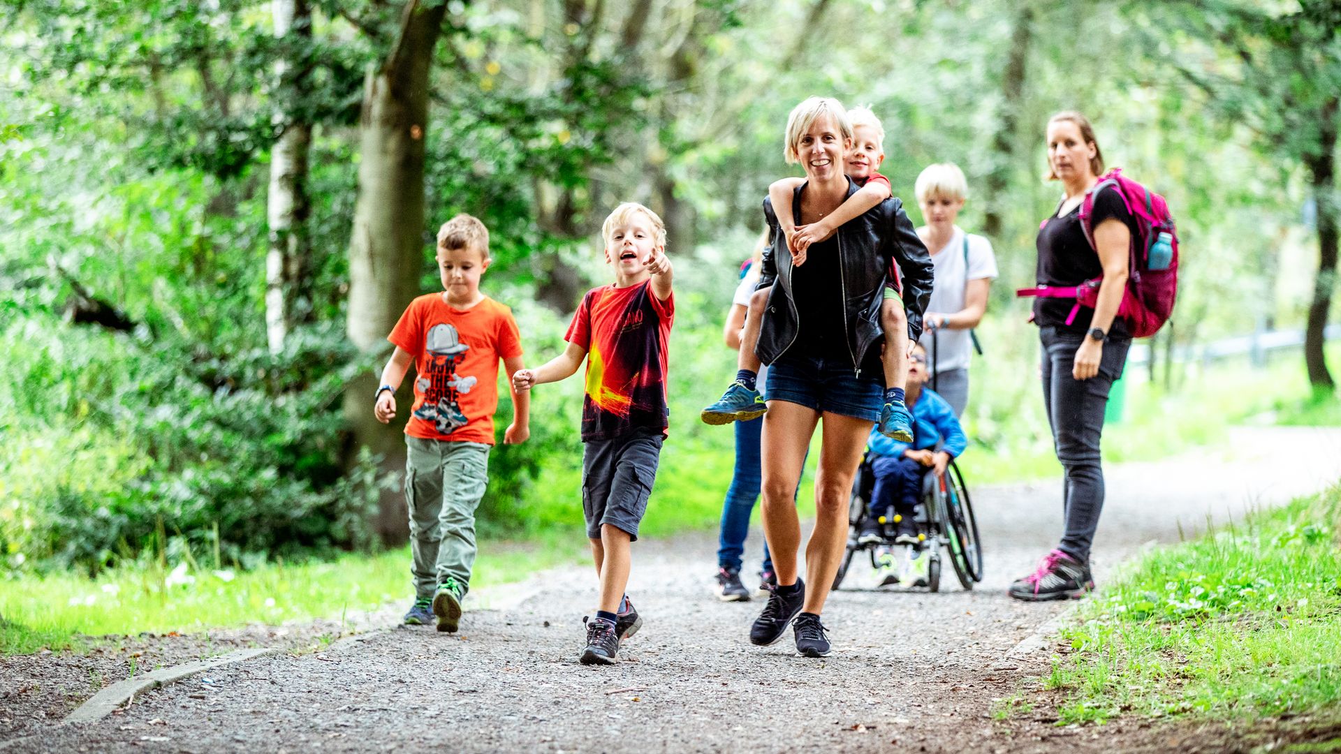 Drei Frauen und fünf Kinder laufen in bunter Sommerkleidung auf den Fotografen zu. Ein Kind sitzt im Rollstuhl und wird geschoben, ein anderes Kind wird von einer Frau auf dem Rücken getragen.