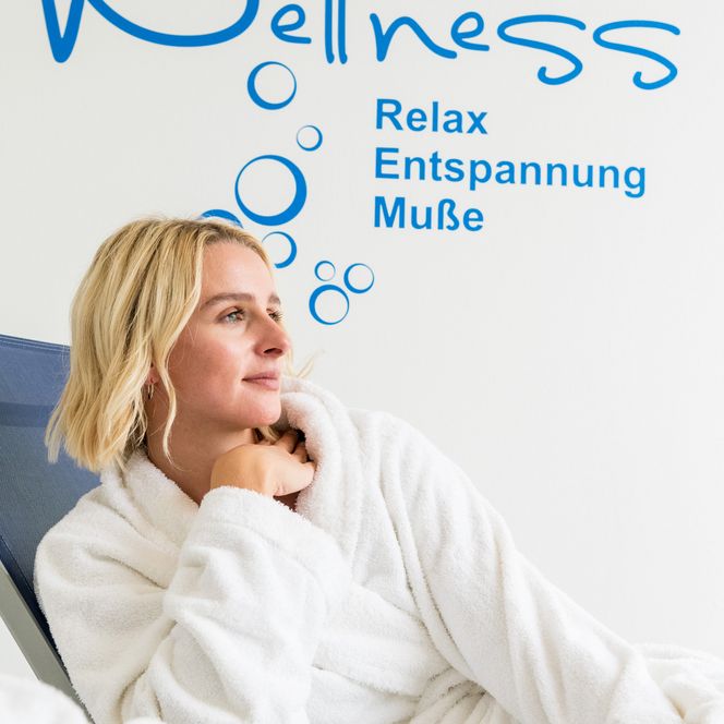 Eine Frau im Bademantel entspannt auf einem Stuhl in einem Wellnessbereich vor einer ruhigen Wand.