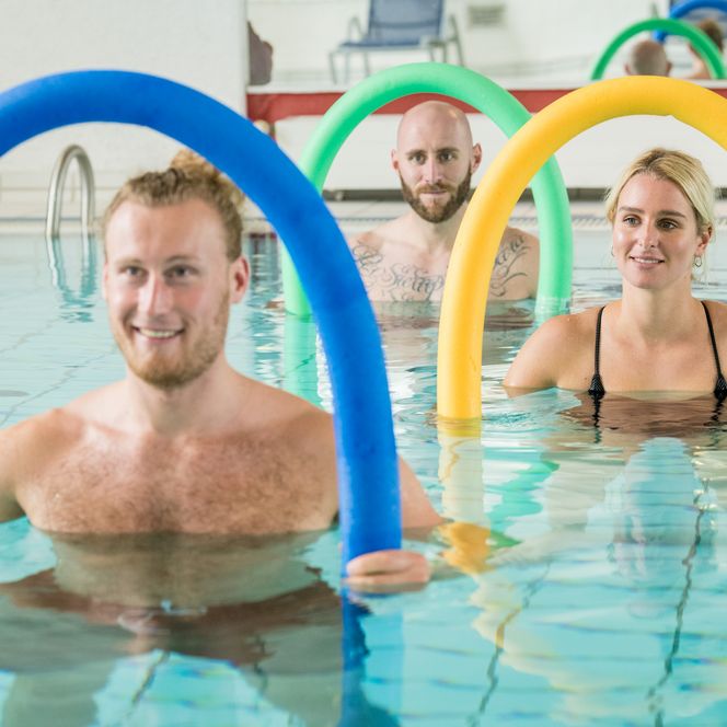 Menschen trainieren lächelnd mit bunten Poolnudeln im Schwimmbad. Das Wasser ist klar und leuchtend.
