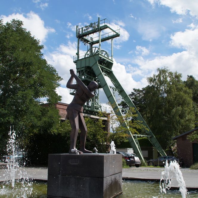Fördertürme und Skulptur eines Bergmanns am Wasserspiel, blauer Himmel, grüne Bäume im Hintergrund.