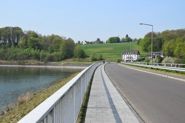 Eine Landstraße führt entlang eines Staudammes, umgeben von grünen Feldern und Wäldern.
