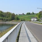 Eine Landstraße führt entlang eines Staudammes, umgeben von grünen Feldern und Wäldern.