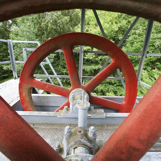 <p>Rotes Metallrad eines alten Wasserversorgungssystems vor grünen Bäumen, von einer Plattform aus gesehen.</p>