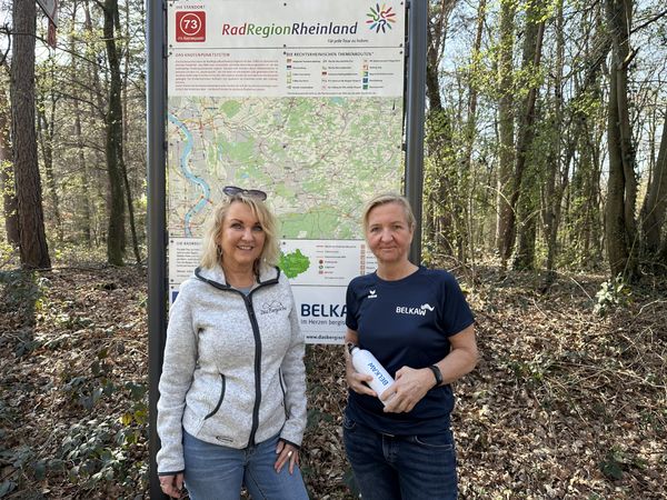 Zwei Frauen stehen vor einer Infotafel im Wald. Eine von ihnen hält eine Trinkflasche in der Hand.