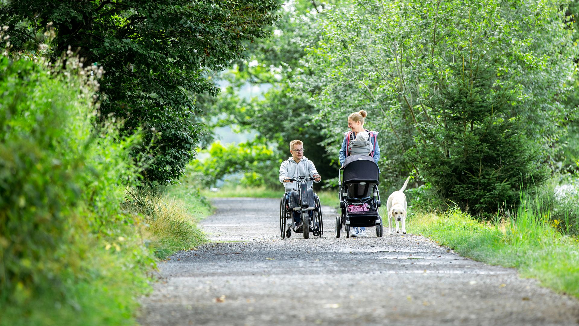 Ein Mann in einem Handbike und eine Frau die einen Kinderwagen schiebt laufen auf einem breiten Wanderweg auf den Fotografen zu. Neben ihnen läuft ein hellbrauner Hund.