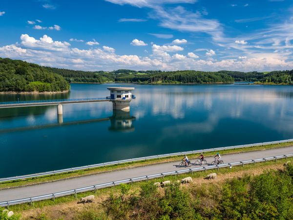 Luftaufnahme von drei Radfahrern, die hintereinander über den Staudamm einer Talsperre fahren. Vor ihnen ist eine Böschung zu sehen, hinter ihnen die Talsperre und ein Turm. 