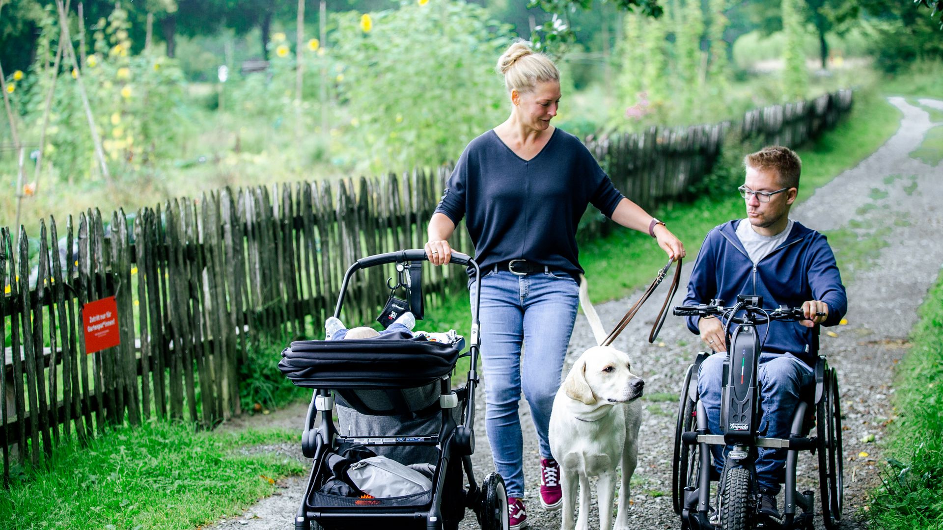 Eine Frau mit Kinderwagen in der rechten Hand und einem Hund an einer Leine in der linken Hand geht über einen Wanderweg. Neben ihr fährt ein Mann mit einem Handbike.