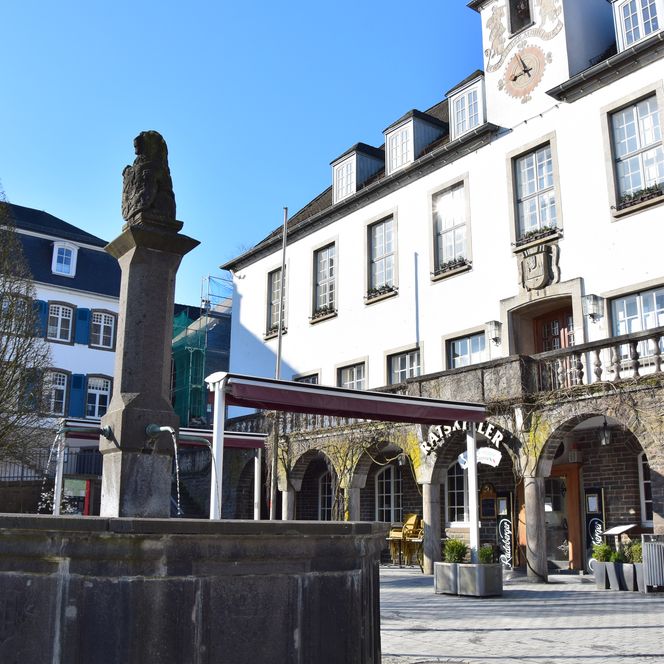 Weißes historisches Gebäude mit Uhrturm und Bogengang neben einem Brunnen. Klarer, blauer Himmel.