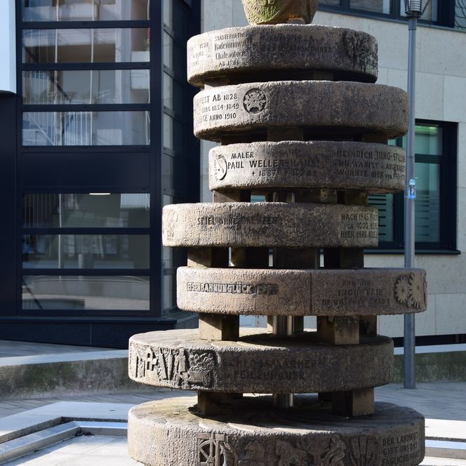 Runde Steinskulptur mit mehreren beschrifteten Scheiben und Wappenschild an Oberseite.