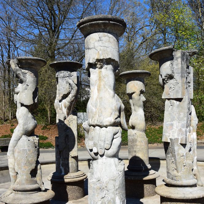 Säulen mit abstrakten Skulpturen in einem Park, umgeben von Bäumen unter klarem, blauem Himmel.