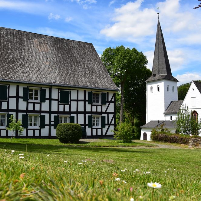 Fachwerkhaus und Kirche in Bergneustadt vor blauem Himmel, umgeben von grünen Bäumen und Wiese.