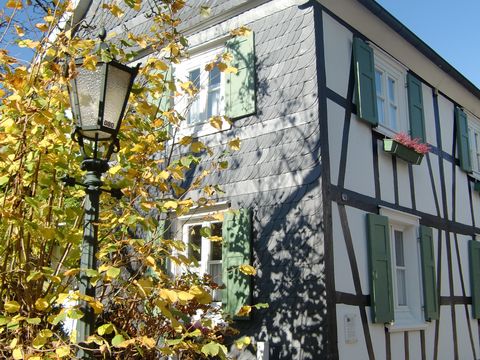 Auf diesem Bild ist ein Fachwerkhaus von der schrägen Seite zu sehen. Rechts sieht man das Fachwerk, und die Linke Seite zeigt die Schieferwand des Hauses. Links neben dem Haus ist ein gelber Baum zu sehen, vor dem eine alte Laterne steht.