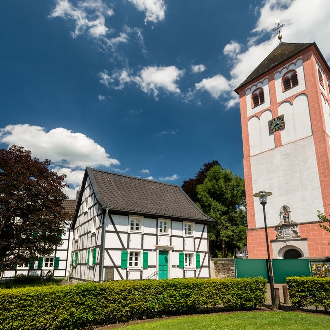 St. Pankratius Kirche in Odenthal: Fachwerkhaus und Kirchturm bei sonnigem Wetter, umgeben von Bäumen.