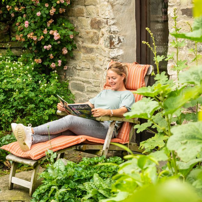 <p>Frau entspannt auf einer Sonnenliege vor einem steinernen Haus, umgeben von blühenden Pflanzen.</p>