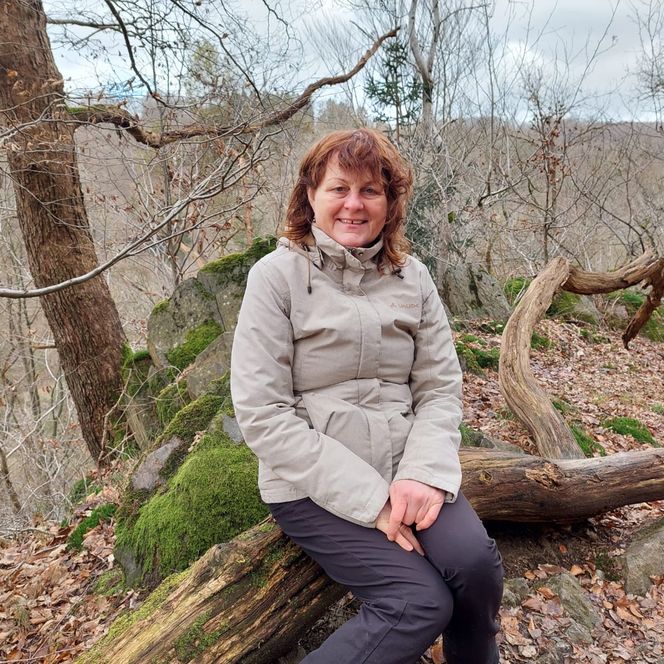 Die Gästeführerin Heide Neumann sitzt auf einem liegenden Baumstamm im Wald.