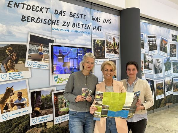 Drei Frauen stehen vor einer beklebten Schaufensterfront eines leeren Ladenlokals in einem Einkaufszentrum. Sie halten gemeinsam einen Flyer in der Hand.