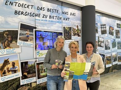 Drei Frauen stehen vor einer beklebten Schaufensterfront eines leeren Ladenlokals in einem Einkaufszentrum. Sie halten gemeinsam einen Flyer in der Hand.