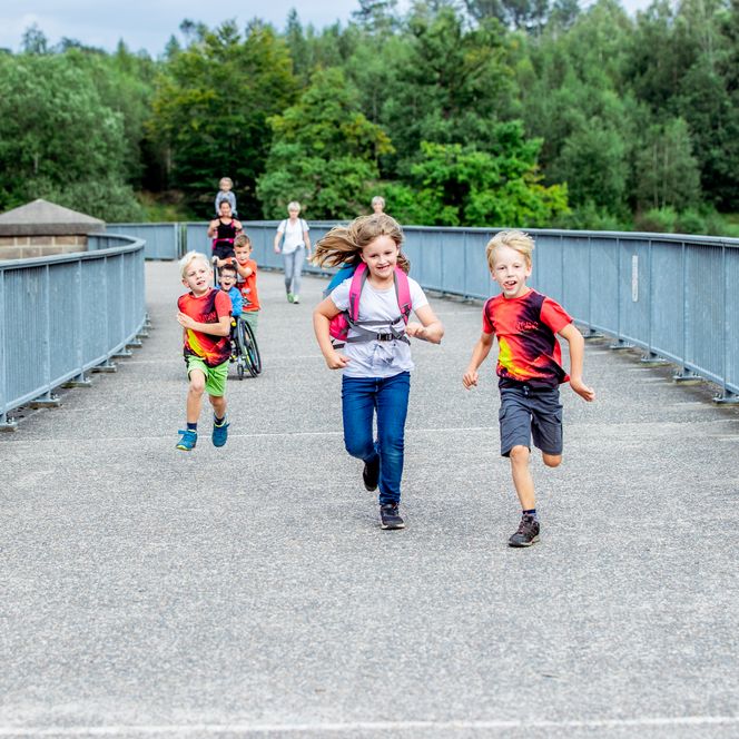 Kinder rennen auf einer Brücke, umgeben von Wald und Natur, voller Freude und Energie.