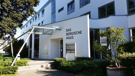 Moderne Gebäudefront mit großem Schriftzug "Das Bergische Haus" und angrenzenden grünen Pflanzen.