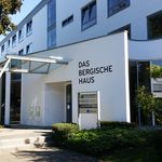 Moderne Gebäudefront mit großem Schriftzug "Das Bergische Haus" und angrenzenden grünen Pflanzen.