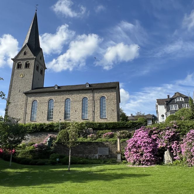 <p>Historische Kirche in Wiehl mit blühenden Rhododendren und blauem Himmel im Hintergrund.</p>
