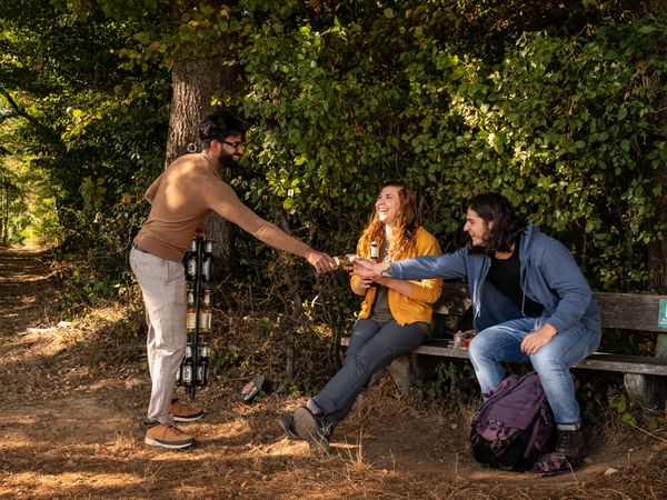 Drei Wandernde treffen sich an einer Bank im Wald; eine stehende Person reicht den beiden Sitzenden ein Getränk.