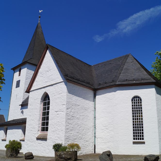 <p>Weiße Kirche in Gummersbach mit steilem, dunklem Dach vor blauem Himmel und grünen Bäumen.</p>